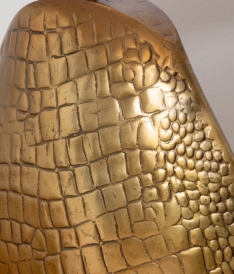 Gros plan sur le relief et la patine dorée du pied de lampe façon croco embossé.