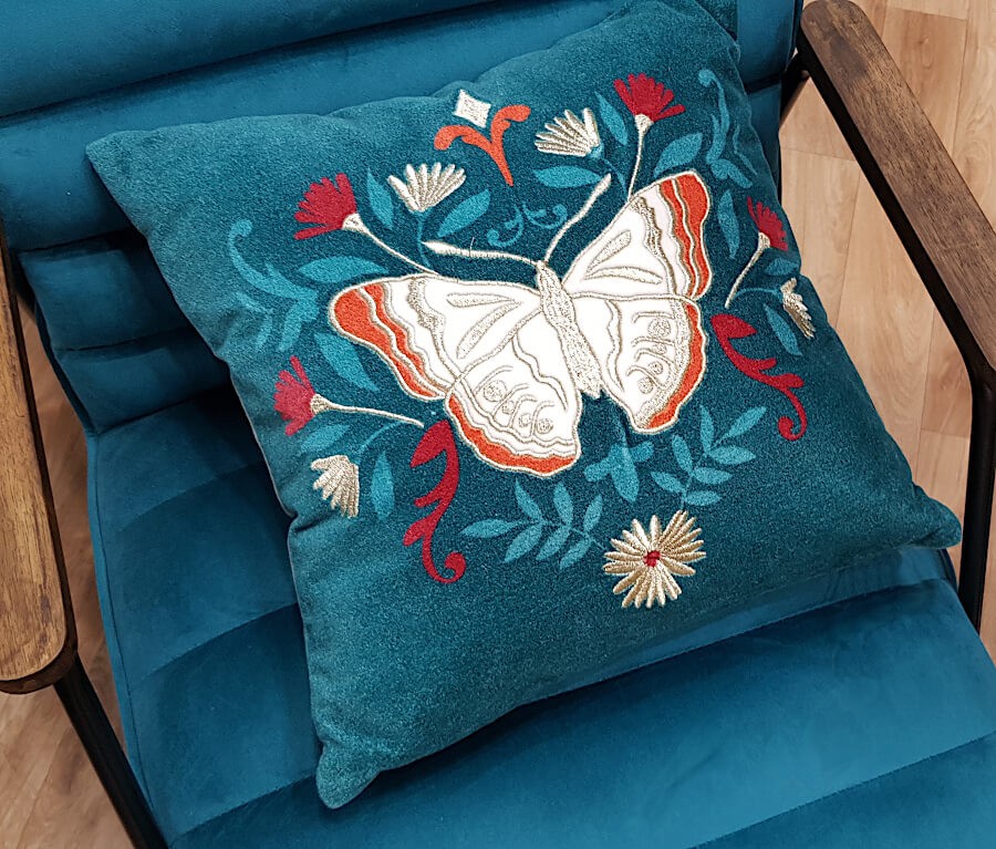 Coussin décoratif papillon bleu canard posé sur un fauteuil en velours bleu.