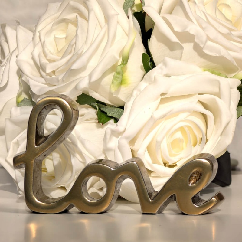 Statuette Love en métal doré posée devant un bouquet de roses blanches artificielles Deco-Delight.