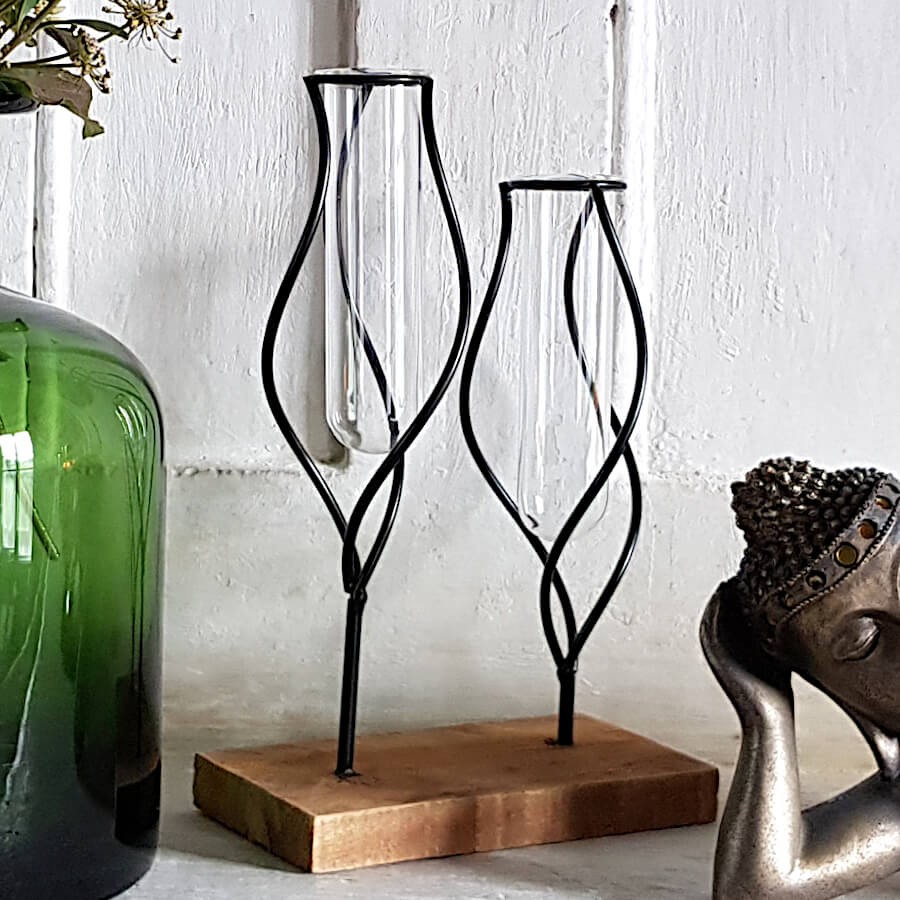 Détail des lignes ondulées en métal noir enserrant les tubes en verre de ce soliflore double posé sur une cheminée en pierre.