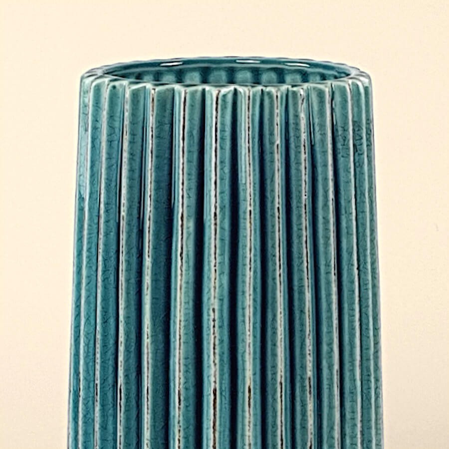 Focus sur le col et les arêtes patinées du vase bleu, sur fond uni doux.