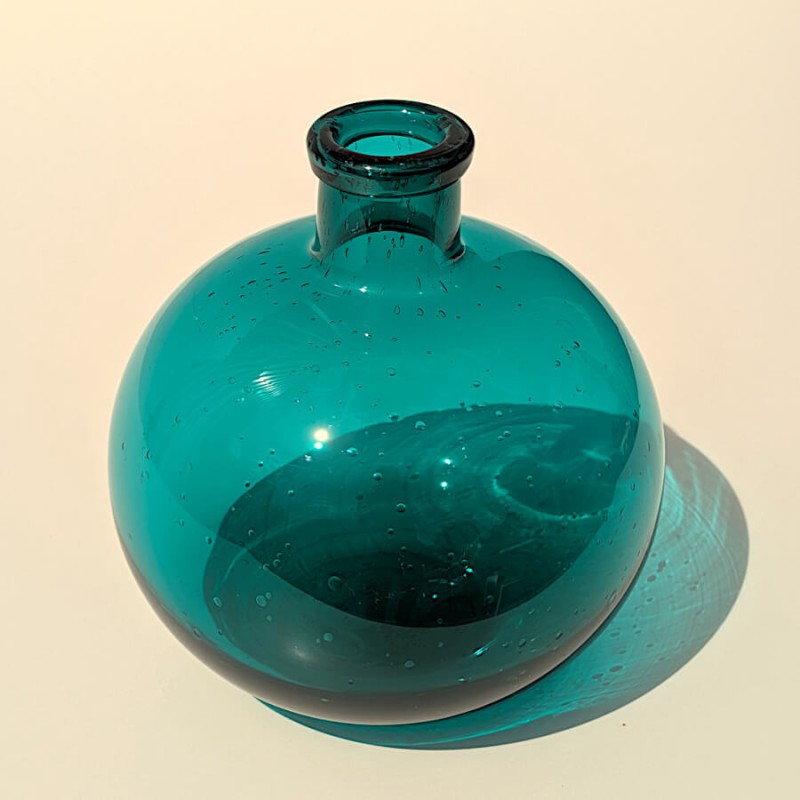 Le vase en verre bullé teinté de turquoise au soleil, créant un jeu d'ombre et de lumière apaisant.