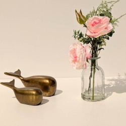 Mise en scène épurée : Le vase fleuri posé à côté de deux statuettes de baleines dorées sur un fond clair.