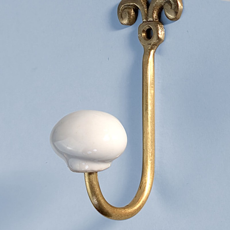 Zoom sur la courbure du crochet en laiton doré et la finition bombée de la boule en céramique blanche sur fond bleu clair.