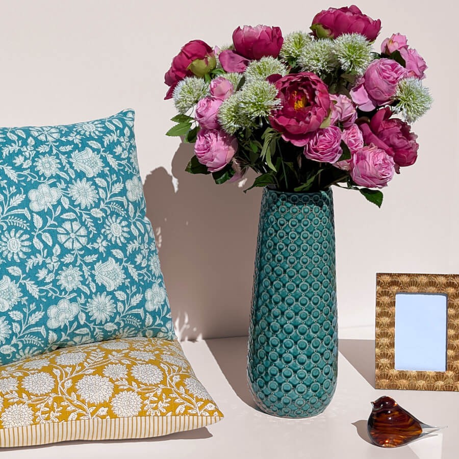 Mise en scène exotique : Vase bleu turquoise rempli de pivoines près de deux coussins fleuris.