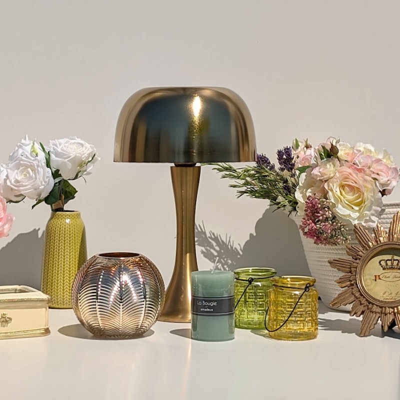 Collection d'objets : Photophore en verre à motif végétal à côté d'une bougie verte et d'une lampe Champignon dorée.