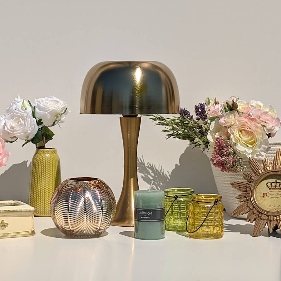 Collection d'objets : Photophore en verre à motif végétal à côté d'une bougie verte et d'une lampe Champignon dorée.
