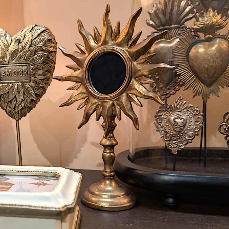 Composition ex-voto : Miroir soleil or mis en scène sur un meuble sombre aux côtés de cœurs sacrés sous cloche.