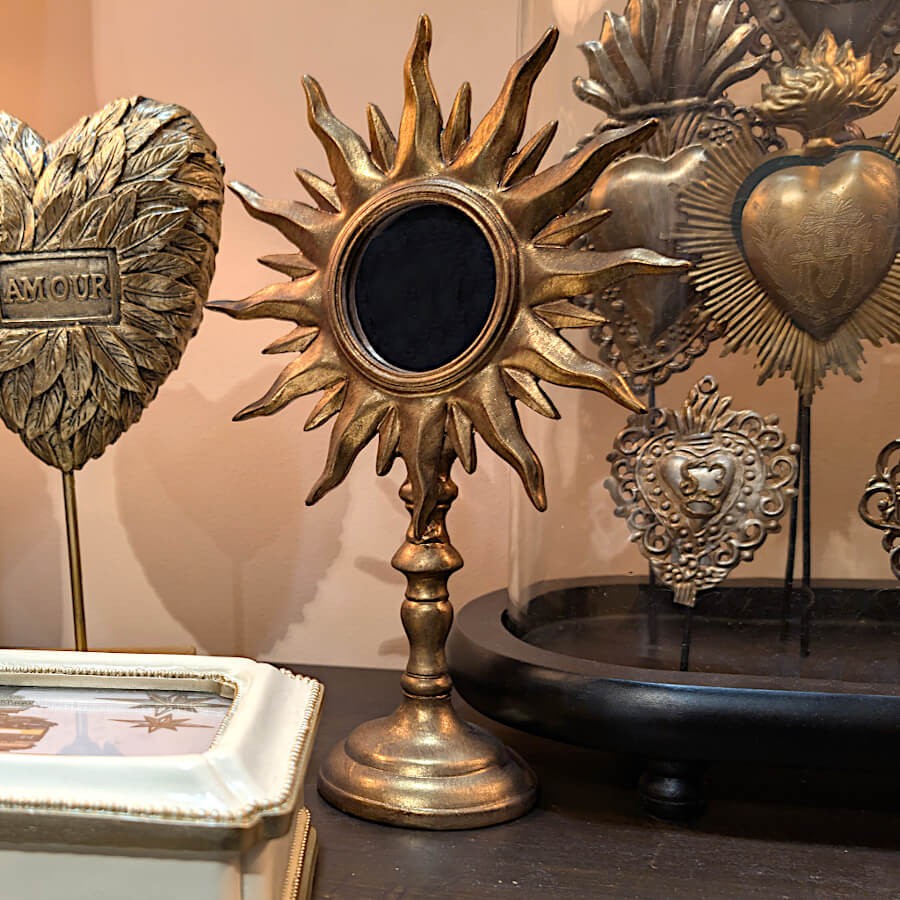 Composition ex-voto : Miroir soleil or mis en scène sur un meuble sombre aux côtés de cœurs sacrés sous cloche.