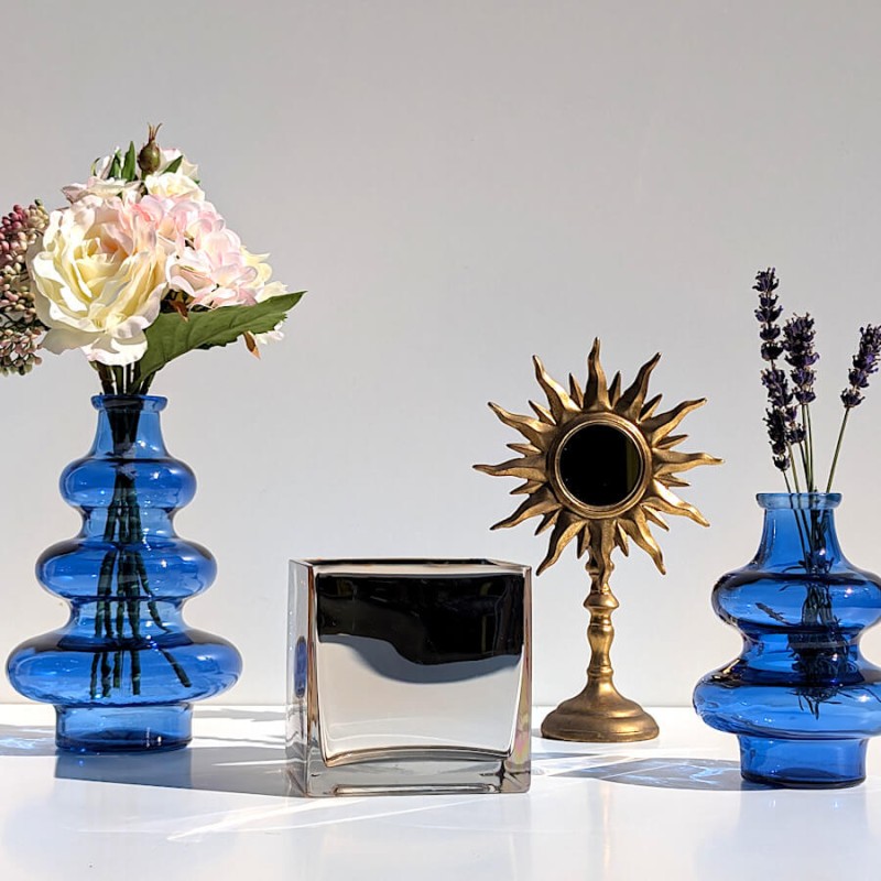 Composition estivale avec le petit cube miroité doré, des vases bleus et un miroir soleil sur pied.