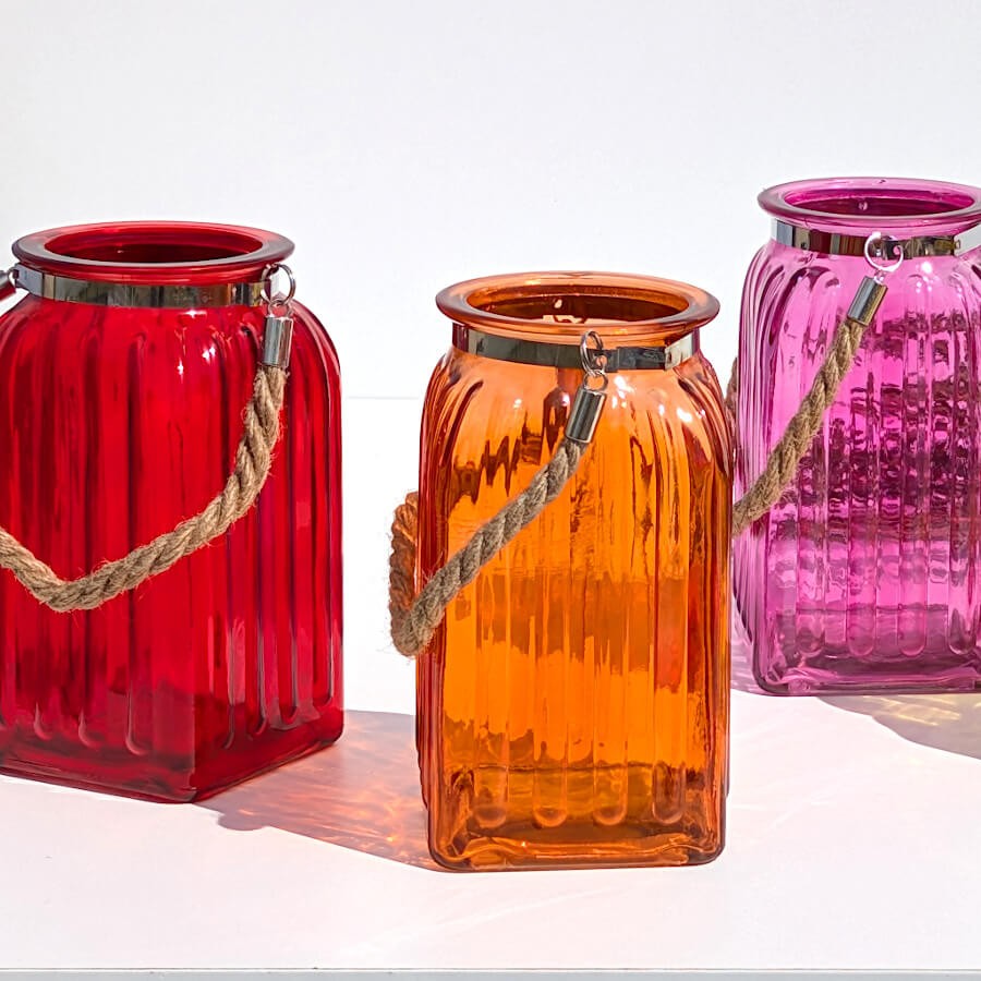 Trio chaleureux : Alignement de photophores en verre teinté rouge, orange et rose fuchsia sur fond blanc.