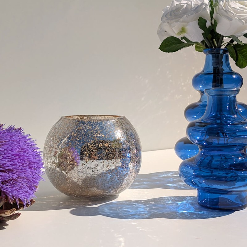 Le photophore craquelé posé à côté d'un grand vase en verre bleu à vagues Deco-Delight sur une surface blanche ensoleillée.