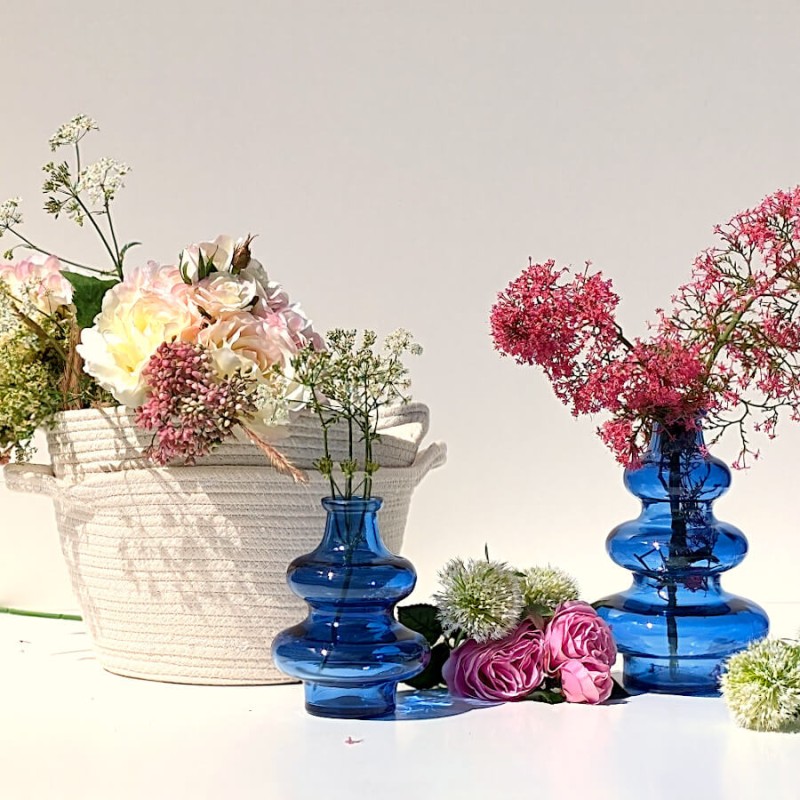 Ambiance printanière : Panier blanc cassé utilisé comme cache-pot pour un grand bouquet de fleurs près de vases bleus.