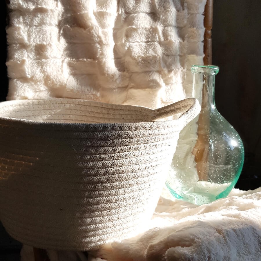 Corbeille en tissu blanc placée sur un plaid en fausse fourrure à côté d'une dame-jeanne en verre. Collection Deco-Delight.