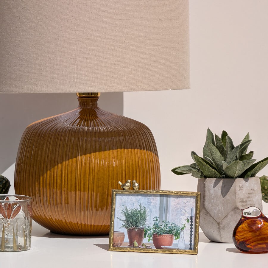 Ambiance chaleureuse avec une lampe à poser en céramique striée ocre, un cadre photo de plantes et le pot facetté gris.
