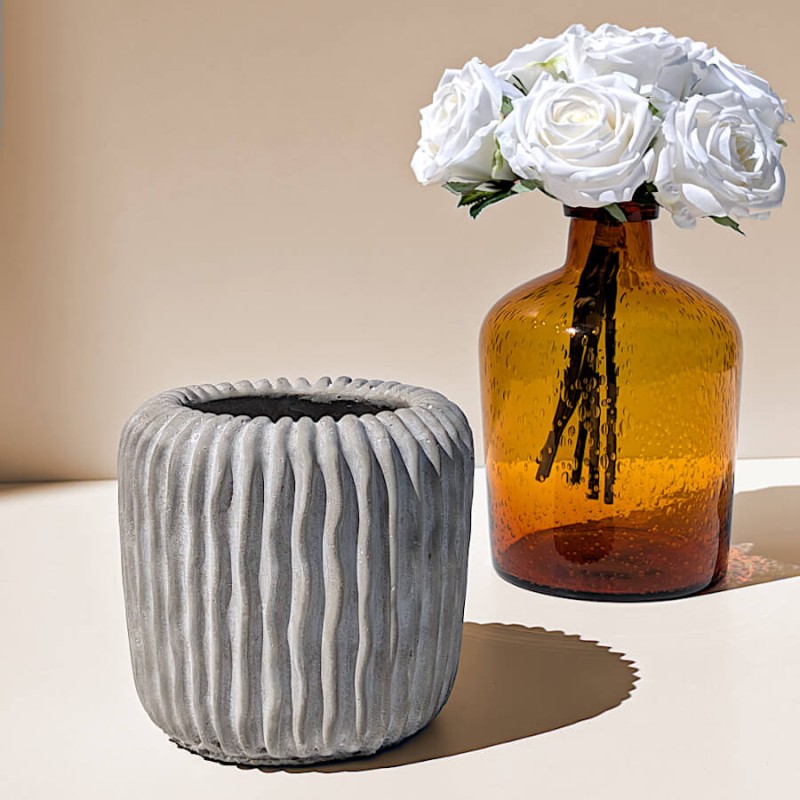 Le pot en ciment côtelé présenté sur une étagère claire à côté d'un vase en verre ambré garni de roses blanches.