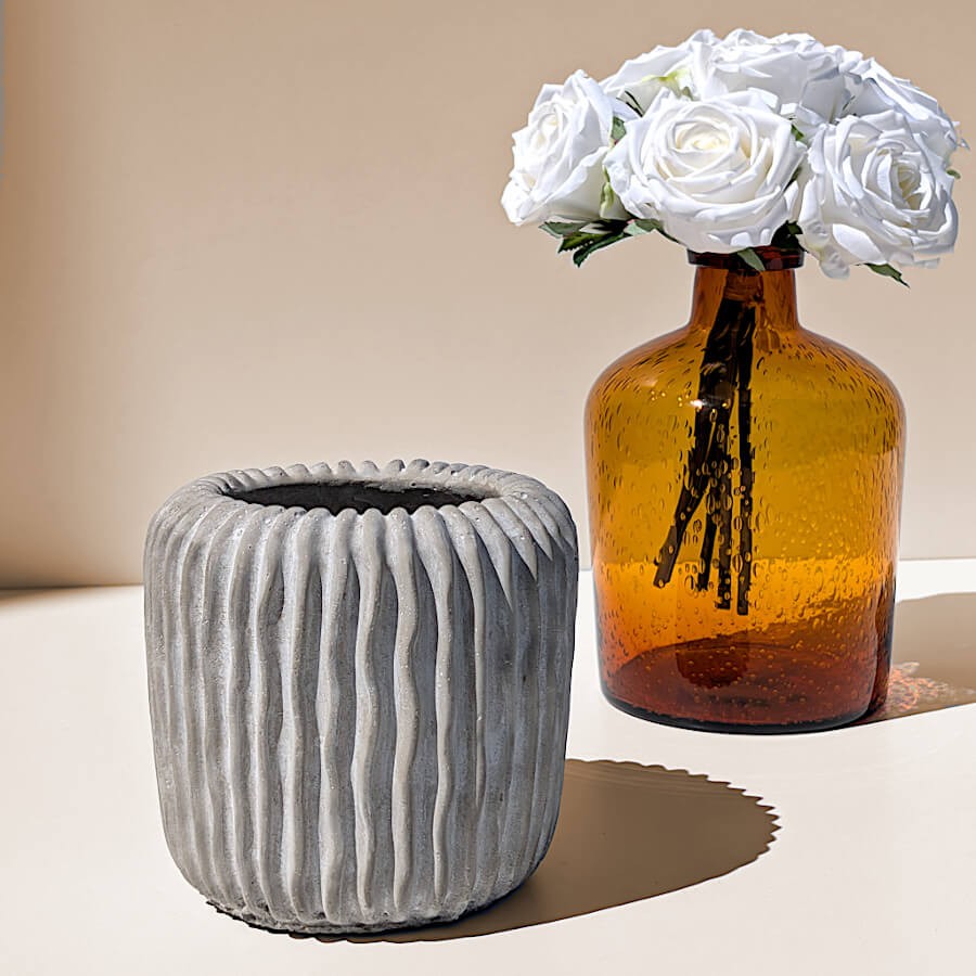 Le pot en ciment côtelé présenté sur une étagère claire à côté d'un vase en verre ambré garni de roses blanches.