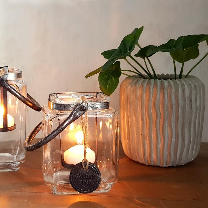 Ambiance de soirée feutrée avec des lanternes en verre et cordage allumées à côté du pot côtelé végétalisé.