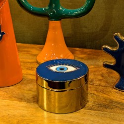 Boîte dorée couvercle œil bleu posée sur meuble en bois à côté d'un bougeoir design.