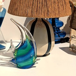 Statuette poisson en verre bleu posée sur un meuble à côté de notre lampe à abat-jour en raphia.
