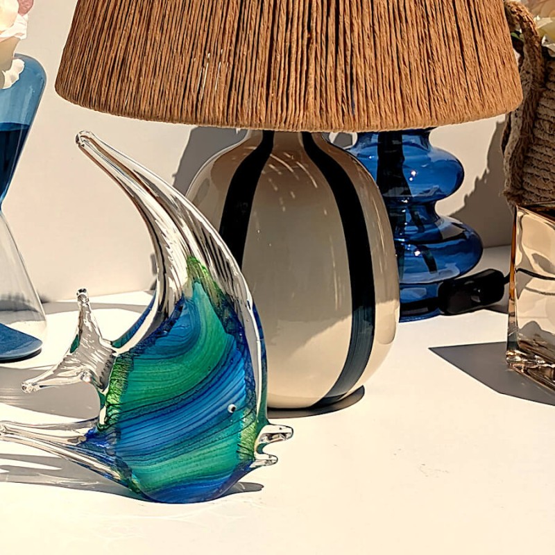 Statuette poisson en verre bleu posée sur un meuble à côté de notre lampe à abat-jour en raphia.
