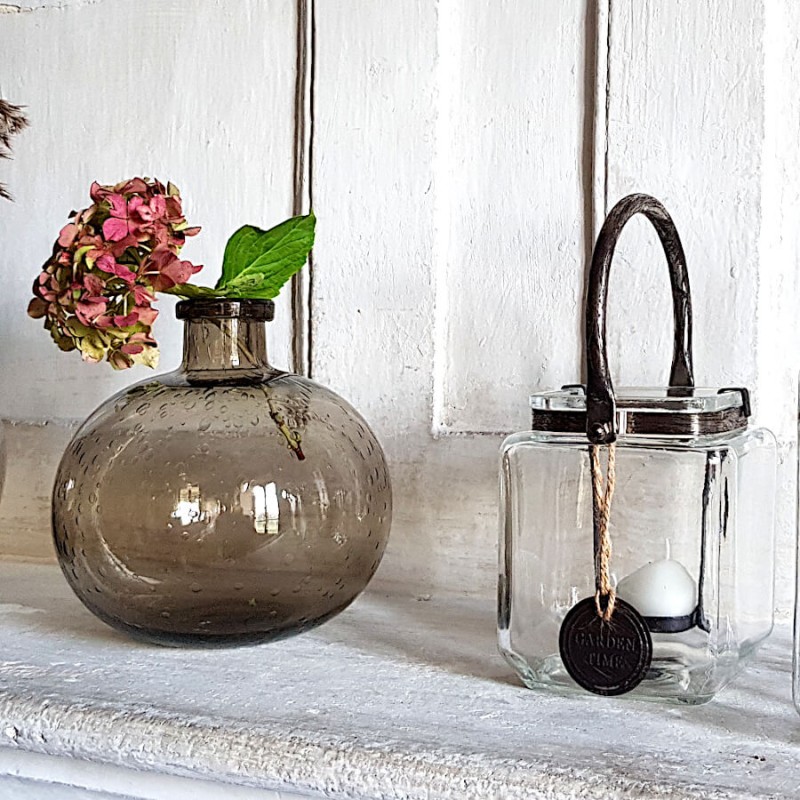 Photophore en verre et métal posé sur un manteau de cheminée à côté d'un vase en verre bullé à la teinte fumée.