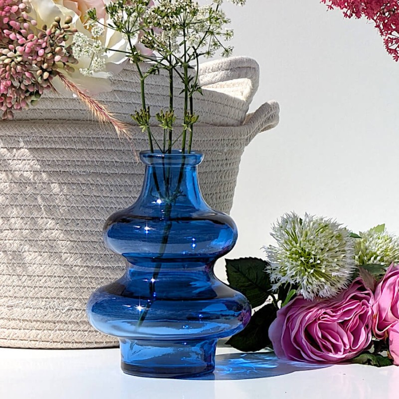 Vase en verre bleu 2 boules garni de petites fleurs blanches posé sur un meuble devant un panier fleuri.