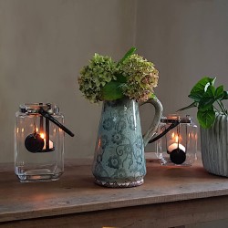 Lanterne carrée en verre et métal patiné posée sur table en bois à côté d'un pichet rustique émaillé.
