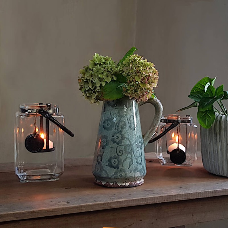 Lanterne carrée en verre et métal patiné posée sur table en bois à côté d'un pichet rustique émaillé.