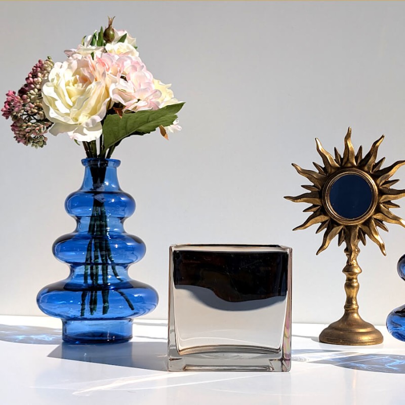 Composition déco avec vase en verre bleu 3 boules fleuri, photophore carré et miroir soleil doré.