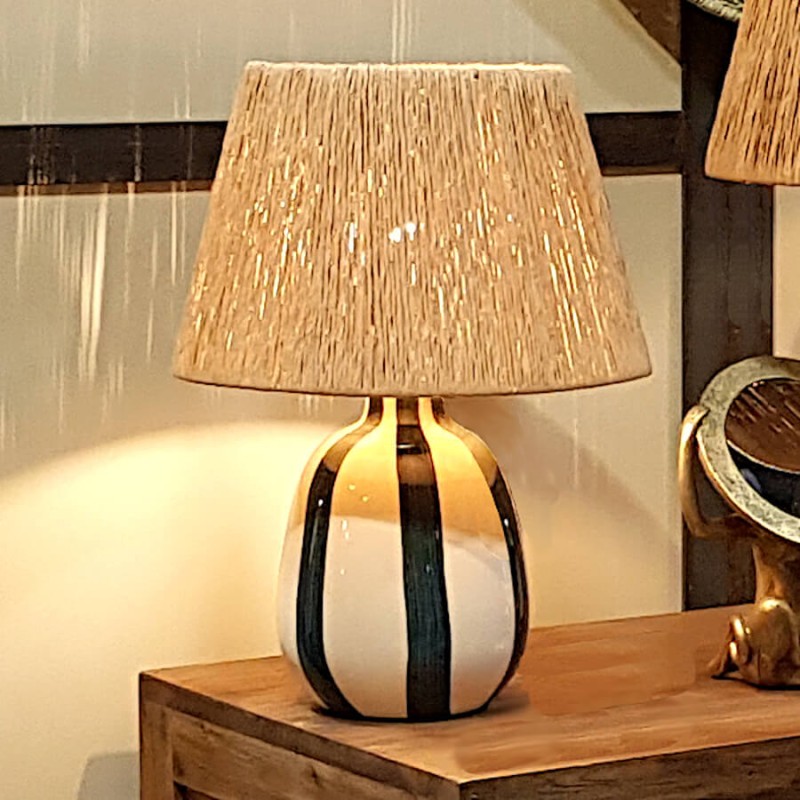 Lampe à poser Riviera allumée sur meuble en bois clair.