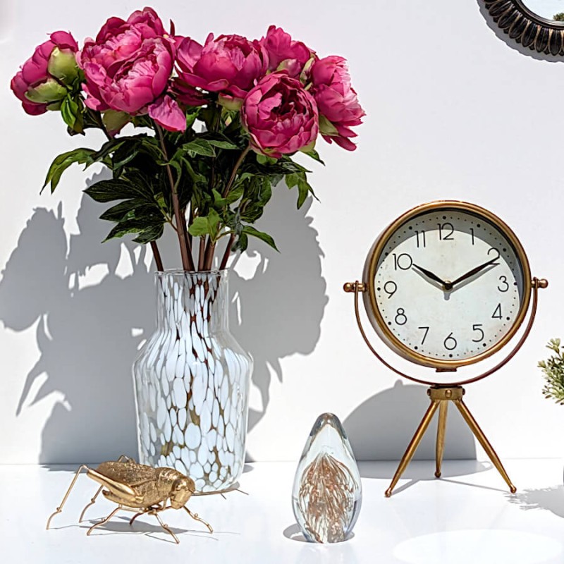 Ambiance bucolique avec le vase garni de pivoines fuchsia à côté de l'horloge rétro dorée et d'une statuette sauterelle dorée.