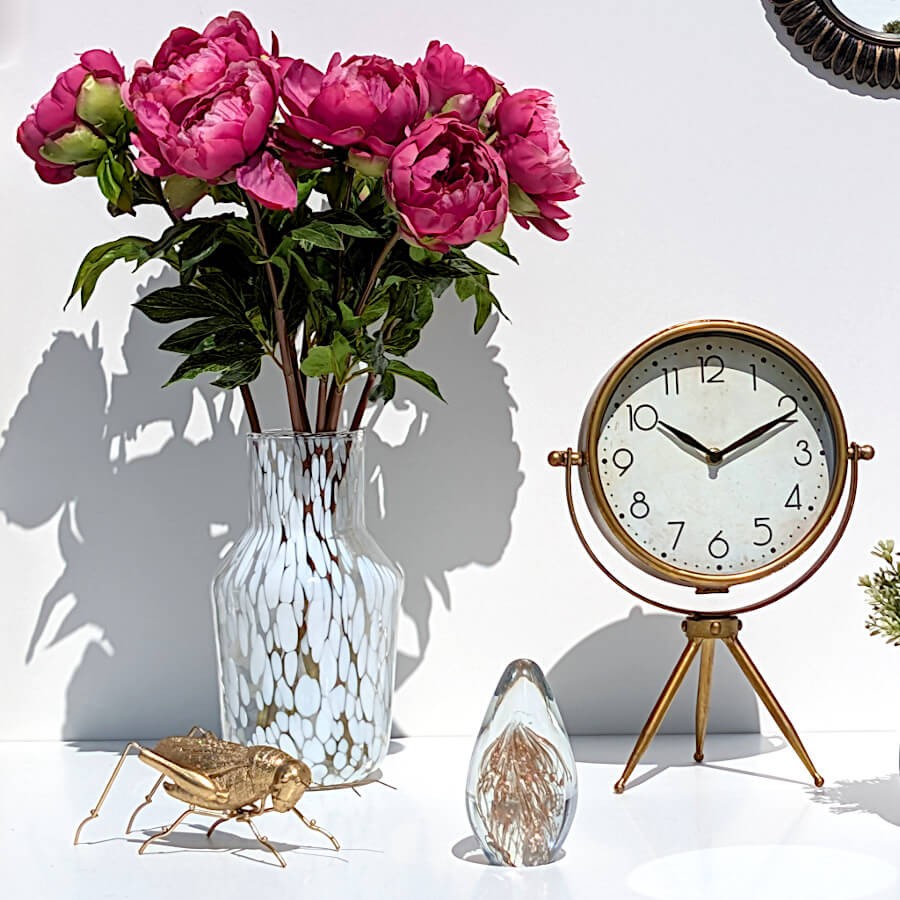Ambiance bucolique avec le vase garni de pivoines fuchsia à côté de l'horloge rétro dorée et d'une statuette sauterelle dorée.
