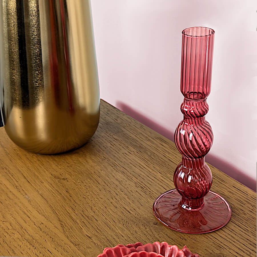 Bougeoir en verre teinté rose framboise présenté à côté d'un pied de lampe à poser doré.